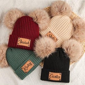 May include: A selection of knitted beanies in shades of burgundy, green, beige, and black. Each hat has a faux fur pom-pom and a leather name patch. The names "Isaiah", "Kaley", "Stella", and "Carter" are displayed on the patches.
