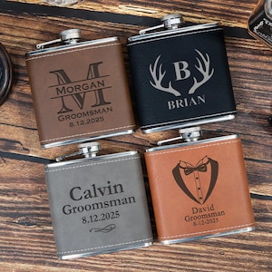 May include: Four personalised hip flasks with silver tops and leather-like exteriors. The flasks are brown, black, grey, and tan. Each flask has a name and date engraved. The brown flask says "Morgan Groomsman 8.12.2025".