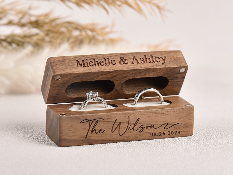 Puede incluir: Una caja de anillos de madera con dos compartimentos para anillos. La caja est&aacute; grabada con los nombres "Michelle & Ashley" y "The Wilson" con la fecha "08.26.2024".