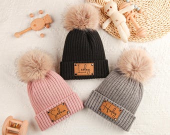 Può includere: Tre berretti a coste in nero, rosa e grigio, ciascuno sormontato da un soffice pompon. I berretti hanno una toppa in pelle con un nome. Accessori per bambini sparsi intorno ai berretti.