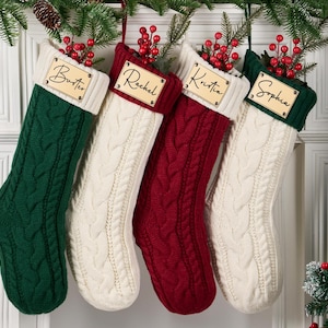 Peut inclure: Quatre bas de Noël en tricot torsadé, verts, crème et rouges, chacun avec une étiquette nominative en bois. Les bas sont accrochés à une porte blanche, décorée de verdure et de baies, créant une scène de vacances festive.