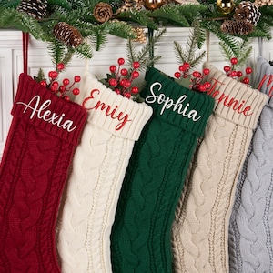May include: Five cable-knit Christmas stockings in red, cream, green, beige, and grey, each with a name embroidered in red. The stockings are decorated with red berries and greenery, hanging from a white mantelpiece.