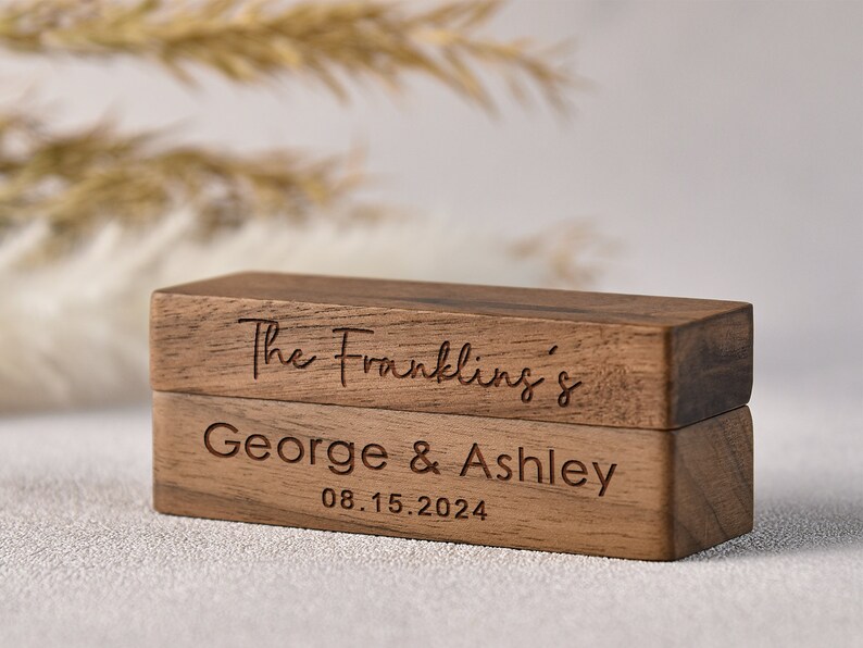 Puede incluir: Una caja de anillos de madera de dos niveles. La capa superior est&aacute; grabada con "The Franklins'" y la capa inferior est&aacute; grabada con "George & Ashley 08.15.2024".