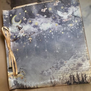 May include: A decorative journal with a celestial theme. The cover features a night sky with stars, clouds, and owls. One owl wears a witch hat and rides a broom. A gold ribbon binds the journal.