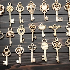 May include: A collection of gold-colored decorative keys with ornate designs, including heart, wing, and floral motifs. The keys are arranged on a dark wooden surface, showcasing their intricate details and vintage aesthetic. Ideal for crafting or jewelry making.