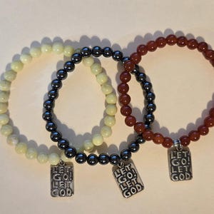 May include: Three beaded bracelets with silver charms that say "Let Go! Let God". One bracelet is made of light green beads, one is made of black beads, and one is made of red beads.