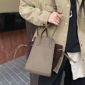 K&ouml;nnte beinhalten: Eine hellbraune Ledertasche mit braunem Wildlederboden und einem langen braunen Riemen. Die Tasche hat zwei obere Henkel und einen Kordelverschluss.