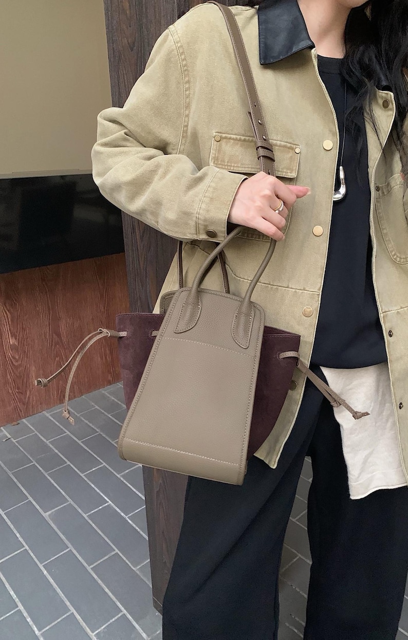 K&ouml;nnte beinhalten: Eine hellbraune Ledertasche mit braunem Wildlederboden und einem langen braunen Riemen. Die Tasche hat zwei obere Henkel und einen Kordelverschluss.
