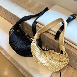 May include: Two mesh shoulder bags. One is black with a black strap, and the other is beige with a beige strap and black leather accents. Both bags have a rounded shape and are displayed on a tiled surface.