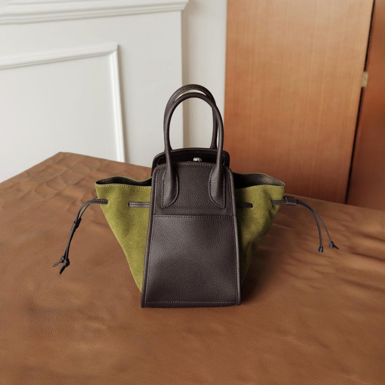 K&ouml;nnte beinhalten: Eine braune Ledertasche mit gr&uuml;nem Wildlederboden und zwei langen Henkeln. Die Tasche hat einen Rei&szlig;verschluss oben und eine kleine Tasche vorne.