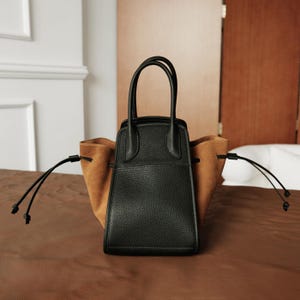 K&ouml;nnte beinhalten: Eine schwarze Ledertasche mit braunen Wildlederakzenten und zwei Kordeln. Die Tasche hat zwei obere Henkel.