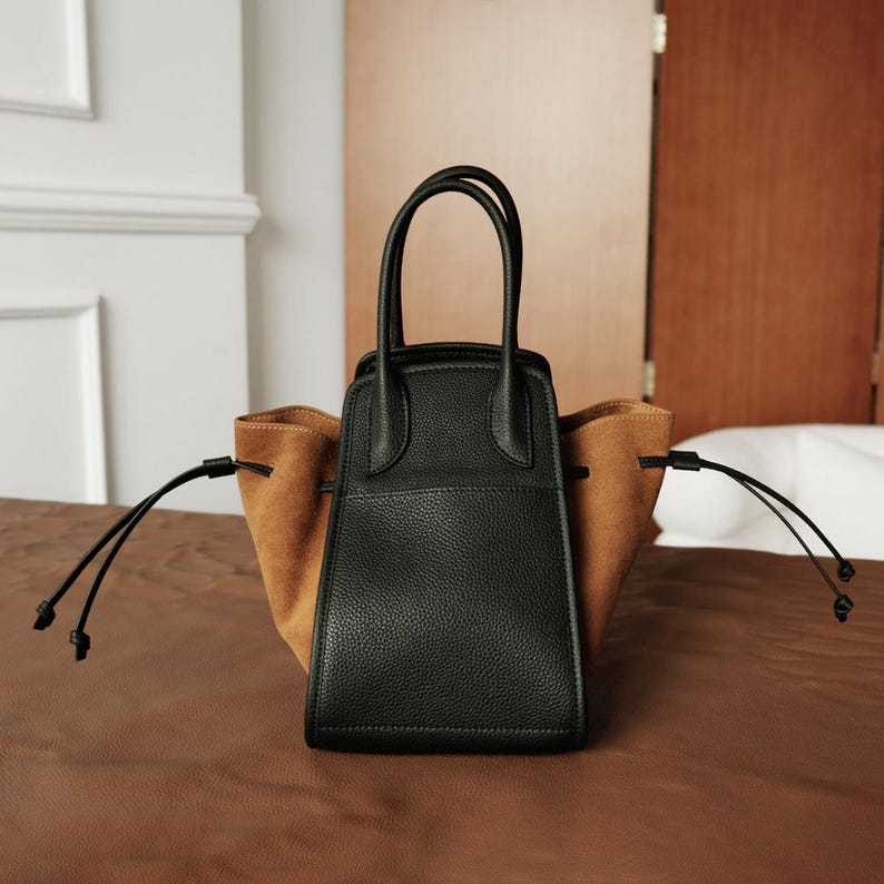 K&ouml;nnte beinhalten: Eine schwarze Ledertasche mit braunen Wildlederakzenten und zwei Kordeln. Die Tasche hat zwei obere Henkel.