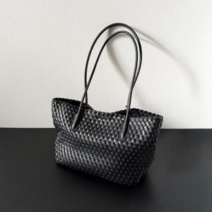 May include: A black woven leather tote bag with two long handles. The bag has a rectangular shape with a woven pattern. The handles are thin and curved, and the bag is sitting on a black surface.