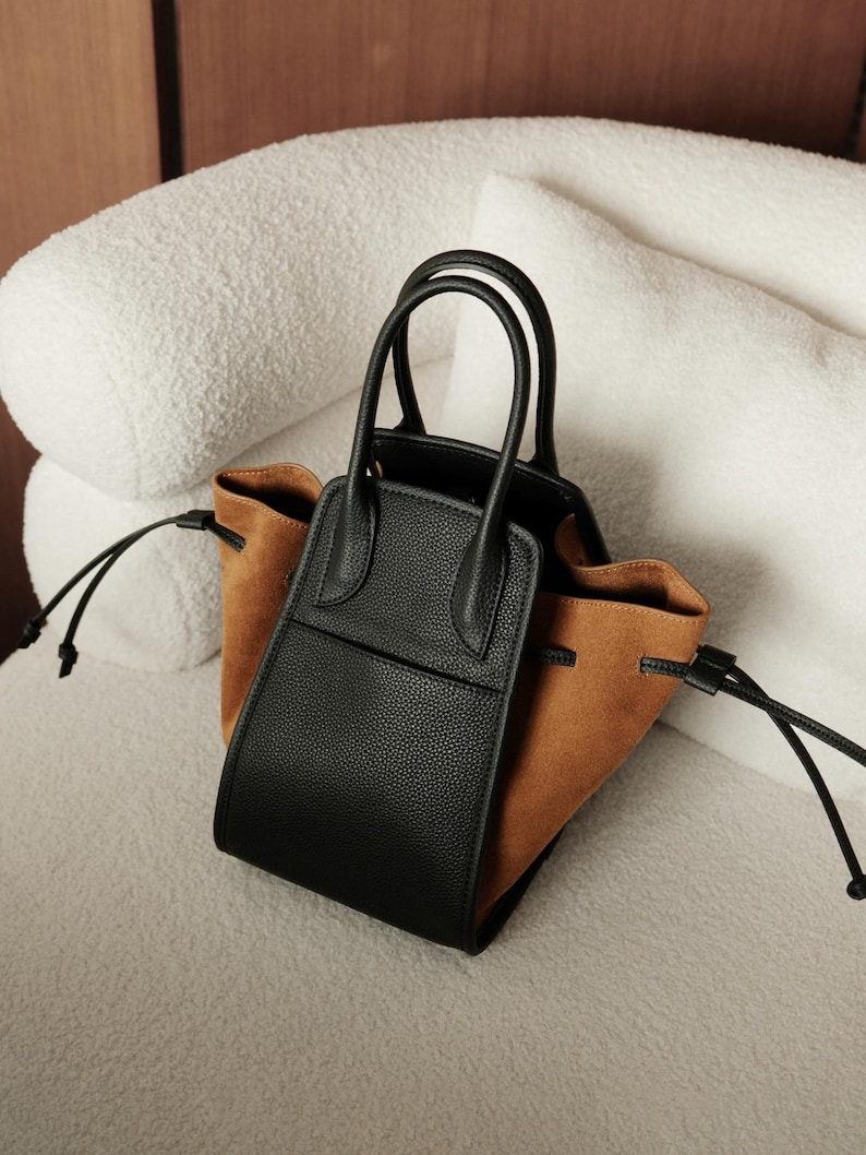 K&ouml;nnte beinhalten: Eine schwarze und braune Ledertasche mit zwei Henkeln oben und einem Kordelzug.