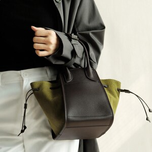K&ouml;nnte beinhalten: Eine braune und olivgr&uuml;ne Ledertasche mit zwei Henkeln oben und einem Kordelzug.