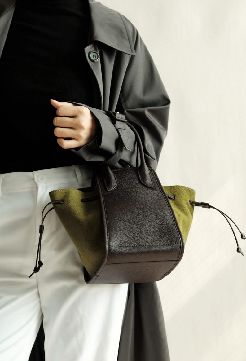 K&ouml;nnte beinhalten: Eine braune und olivgr&uuml;ne Ledertasche mit zwei Henkeln oben und einem Kordelzug.
