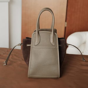 K&ouml;nnte beinhalten: Eine kleine, hellgraue Ledertasche mit einem braunen Wildleder-Einsatz auf der Vorderseite. Die Tasche hat zwei obere Henkel und einen Kordelverschluss.