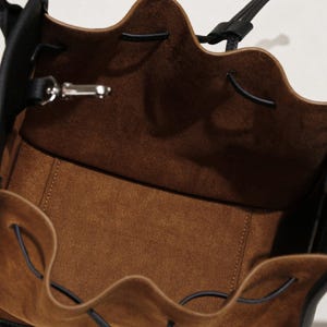 K&ouml;nnte beinhalten: Eine schwarze Ledereimer-Tasche mit braunem Wildlederfutter und einem Kordelzug.