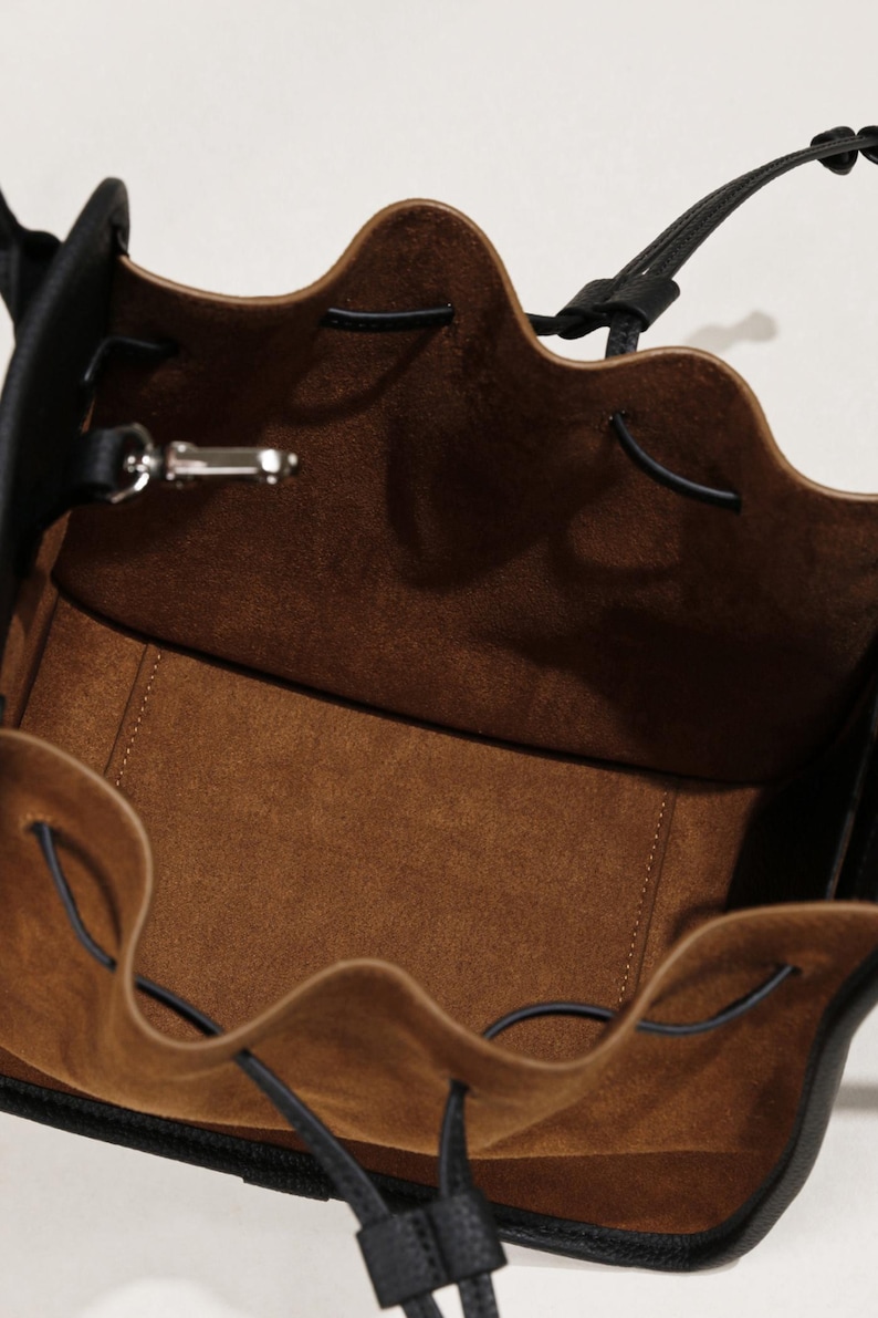 K&ouml;nnte beinhalten: Eine schwarze Ledereimer-Tasche mit braunem Wildlederfutter und einem Kordelzug.