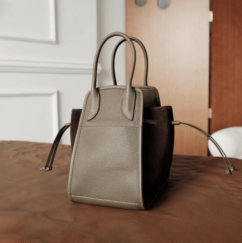 K&ouml;nnte beinhalten: Eine graue Ledertasche mit braunen Wildlederakzenten und einem oberen Henkel. Die Tasche hat einen Kordelverschluss und eine kleine Tasche auf der Vorderseite.
