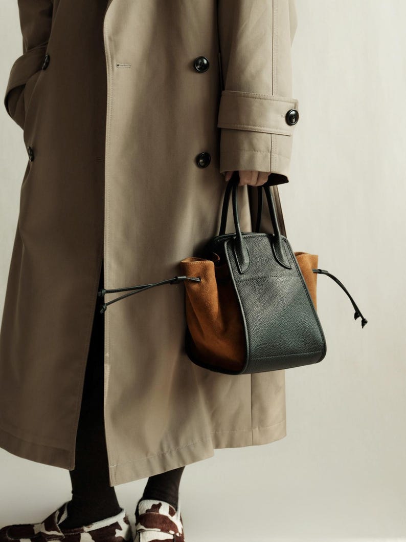 K&ouml;nnte beinhalten: Eine Person tr&auml;gt einen beigefarbenen Trenchcoat und eine schwarze Hose und h&auml;lt eine schwarze und braune Ledertasche mit Kordelzug.