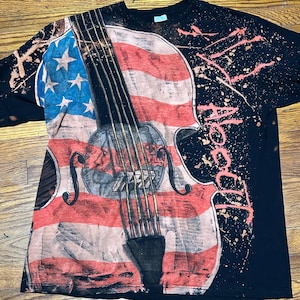Peut inclure: T-shirt noir à manches longues avec un motif vieilli de violon intégrant le drapeau américain. Le t-shirt présente un effet de peinture éclaboussée et le mot "About" en rose.