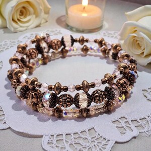 May include: Three-strand bracelet with bronze-colored beads and clear, faceted crystals. The bracelet is set on a white, lacy surface, with a lit candle and white roses in the background. The beads have an ornate, vintage design.