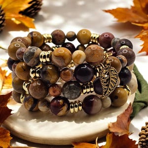 May include: A set of three bracelets made with brown, yellow, and red jasper beads. The bracelets are stacked on top of each other and have gold spacers between the beads. A gold leaf charm hangs from the bottom bracelet.