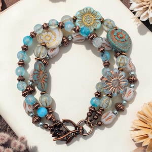 May include: A layered bracelet featuring a mix of blue, white, and copper-toned beads. The bracelet includes flower-shaped and oval-shaped beads, with a decorative clasp. The beads are various sizes and shapes, creating a unique design.