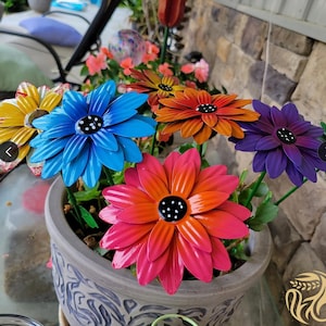 May include: A metal flower arrangement in a gray planter with a floral design. The flowers are painted in various colors, including blue, orange, purple, and red. The flowers are arranged in a circular pattern.