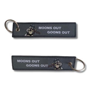 Può includere: Due portachiavi grigi con anelli argentati. Ogni portachiavi presenta il testo bianco "MOONS OUT GOONS OUT", con un'illustrazione di un personaggio che tiene un fucile. I portachiavi sono rettangolari.