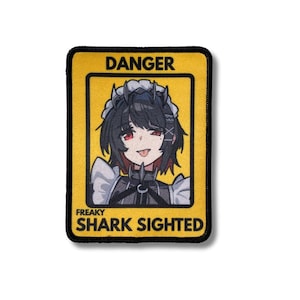 Pode incluir: Patch bordado com fundo amarelo e borda preta. O patch apresenta uma ilustração de desenho animado de um personagem com cabelo preto, olhos vermelhos e uma expressão brincalhona. O texto no patch diz "DANGER" e "FREAKY SHARK SIGHTED."