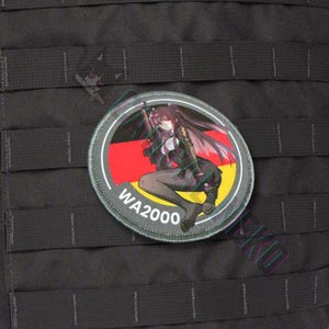 May include: A round patch featuring a manga-style illustration of a woman with long dark hair, holding a rifle. The patch has a German flag design and the text "WA2000". The patch is attached to a black tactical vest.