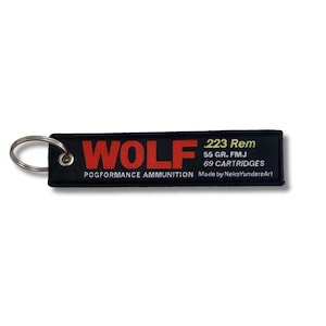 May include: Black rectangular keychain with the word "WOLF" in red block letters. The keychain also includes the text ".223 Rem", "55 GR. FMJ", "69 CARTRIDGES", "POGFORMANCE AMMUNITION", and "Made by NekoYandereArt". A silver key ring is attached.