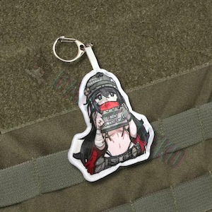 May include: A white patch featuring a cartoon illustration of a female character holding a sign with the text "FRONT TOWARD ENEMY." The character has long black hair, a red scarf, and a military-style hat. A silver keychain is attached.