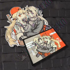 Peut inclure: Deux écussons brodés représentant des illustrations de style anime de deux personnages s'embrassant. Le plus grand écusson est de forme irrégulière, tandis que le plus petit est rectangulaire. Les deux représentent des personnages blonds, aux yeux rouges, en tenue militaire.