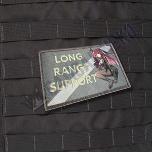 May include: A rectangular patch with the text "LONG RANGE SUPPORT" in gold lettering. The patch features an anime-style illustration of a woman with a rifle. The patch is gray with a black border and is attached to a black tactical vest.