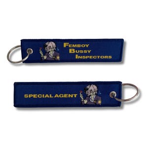May include: Two navy blue keychains with silver key rings. One keychain reads "FEMBOY BUSSY INSPECTORS" and the other reads "SPECIAL AGENT". Both keychains feature a cartoon illustration of a person.