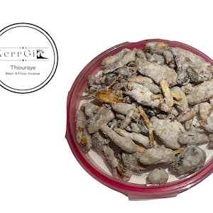 May include: A pink plastic container filled with a mixture of light brown and grey incense sticks. The sticks are covered in a white powder. The container has a label that reads "KerrGi Thiouraye West African Incense".