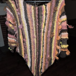 May include: A striped knitted shawl in a variety of colours, including pink, brown, yellow, and white. The shawl has a fringe border and is decorated with small pom-poms.