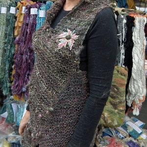 May include: A woman wearing a brown and black knitted vest with a floral brooch. The vest has a unique, textured design. She is standing in front of a table with various knitted items.