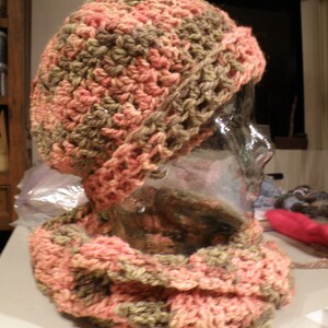 May include: A handmade crochet hat and scarf set. The hat is a beanie style, and the scarf is a cowl. The set is made with a chunky yarn in shades of pink, peach, and olive green. The crochet stitches are visible.