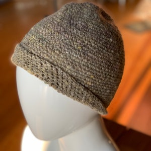 May include: A hand-knitted beanie hat in shades of gray, brown, and green, displayed on a white mannequin head. The hat features a folded brim and a textured, woven appearance, showcasing the craftsmanship of the knit. The hat is a stylish accessory.
