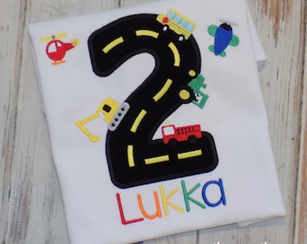 May include: White t-shirt with a large black number 2 design, featuring a road with yellow lines and various vehicles. The name "Lukka" is embroidered in rainbow colors. A red helicopter, yellow excavator, and a blue airplane are also included.