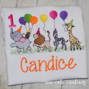 Puede incluir: Una camiseta blanca con un dise&ntilde;o bordado de un n&uacute;mero uno rosa, un elefante gris, un mono marr&oacute;n, un le&oacute;n beige, una cebra blanca y negra y una jirafa, todos con gorros de fiesta y sosteniendo globos de colores sobre hierba verde.