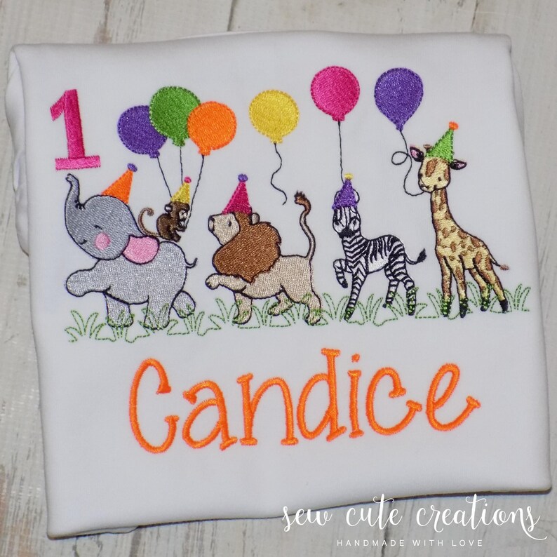 Puede incluir: Una camiseta blanca con un dise&ntilde;o bordado de un n&uacute;mero uno rosa, un elefante gris, un mono marr&oacute;n, un le&oacute;n beige, una cebra blanca y negra y una jirafa, todos con gorros de fiesta y sosteniendo globos de colores sobre hierba verde.