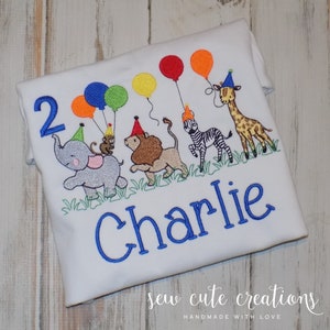 May include: A white t-shirt with blue embroidery of a parade of animals wearing party hats and holding balloons. The text "2" and "Charlie" are also embroidered in blue.