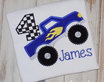 Op de afbeelding: Een wit t-shirt met een geborduurd ontwerp van een blauwe monstertruck met gele vlammen, grote zwarte banden en een geblokt cijfer vier. De naam James is in blauwe schrijfletters onder de truckafbeelding geborduurd.