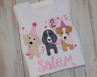 Puede incluir: Una camiseta blanca con un diseño bordado de tres cachorros con gorros de fiesta rosas. El cachorro central sostiene un globo rosa con el número 2. El nombre Salem está bordado en letra cursiva rosa debajo de los perros, rodeado de confeti.