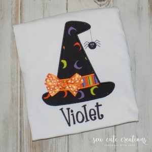 May include: A white t-shirt with an embroidered black witch hat with orange and purple accents, a small orange bow, and a black spider hanging from the hat. The name "Violet" is embroidered below the hat.